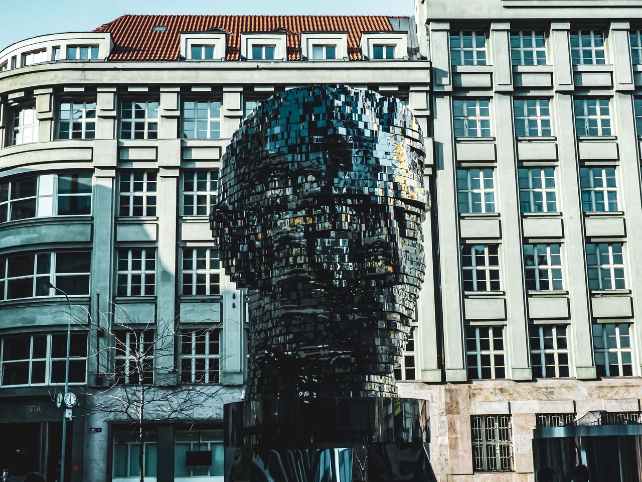 Reflective segmented head sculpture standing in front of a large city building.