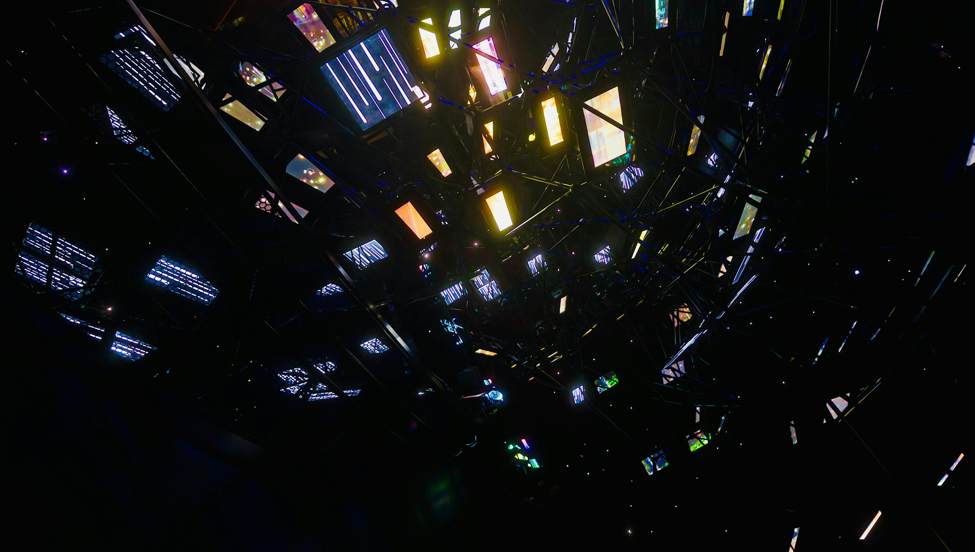 Dark installation filled with suspended illuminated screens and reflective structures scattered through a black space.