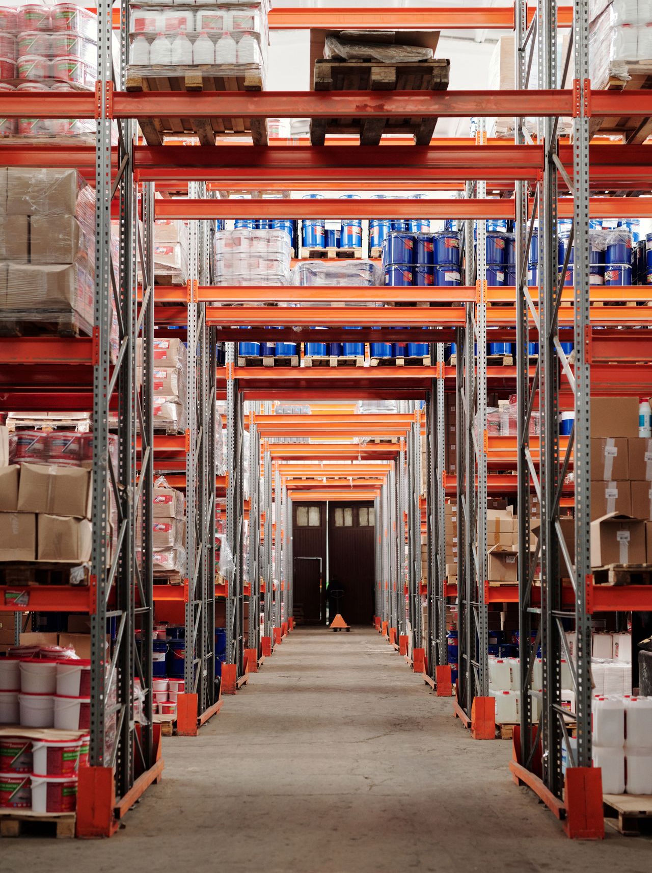 Long warehouse aisle lined with tall storage racks holding pallets, boxes, containers, and drums.