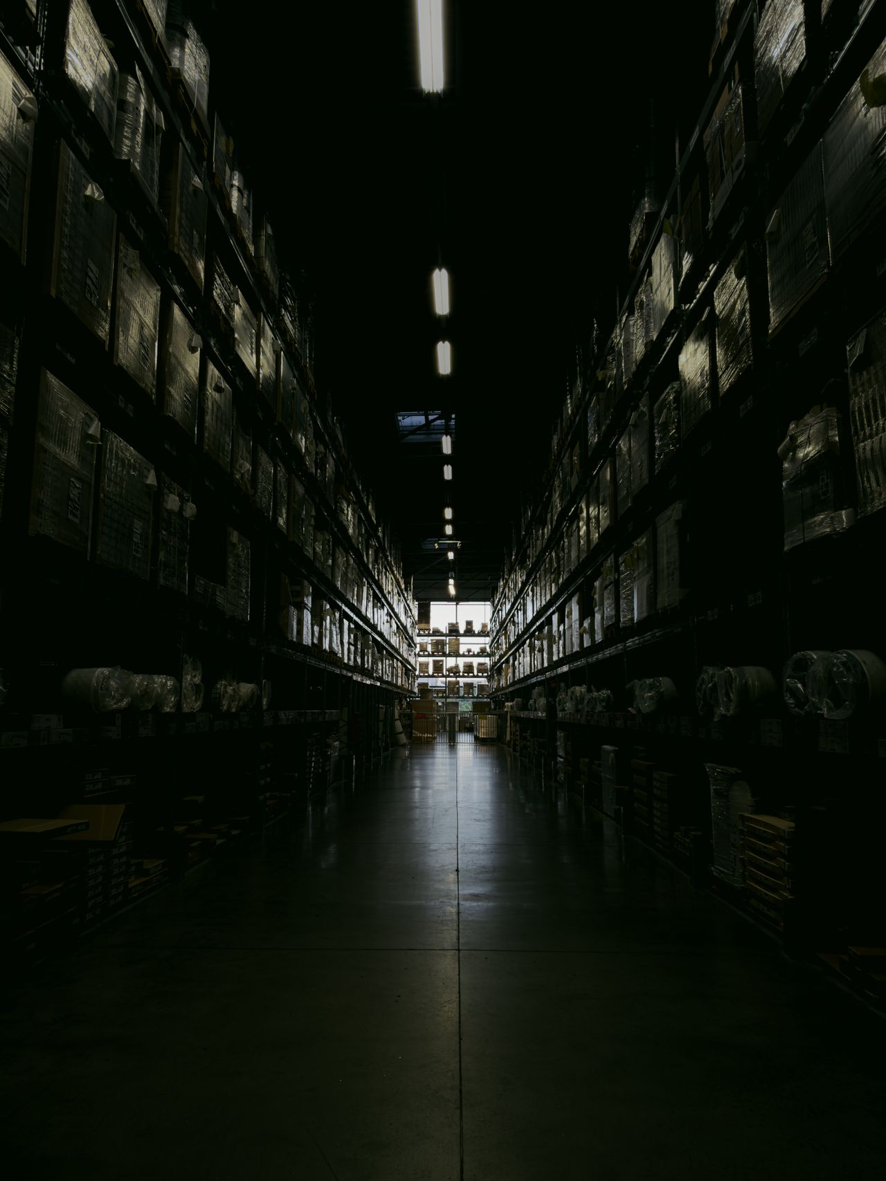 Dark warehouse aisle lined with tall shelves of boxed inventory beneath a row of overhead fluorescent lights.