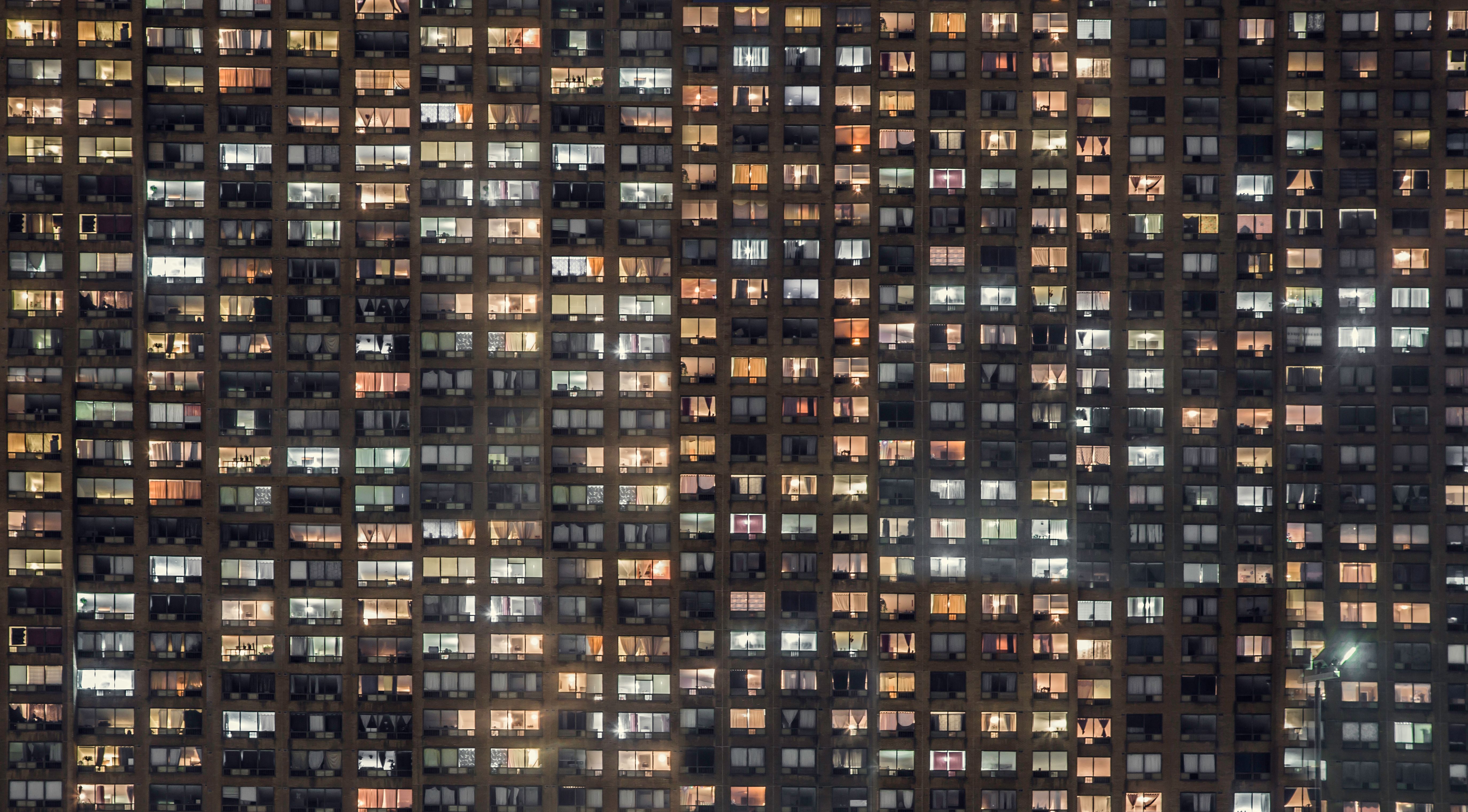 Nighttime facade of a high-rise apartment building with hundreds of illuminated and dark windows arranged in a dense grid.